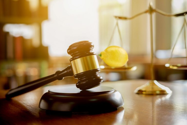 A gavel resting on a judges table. Behind the gavel, there is a scale with a lemon on one side.